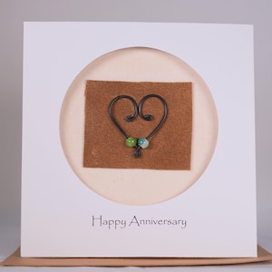 May include: A white card with a brown circle in the center. The circle contains a brown felt square with a black wire heart shape with two green and blue beads. The card says "Happy Anniversary" at the bottom.