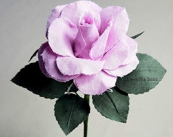 May include: A single lavender fabric rose with green fabric leaves on a stem. The rose is made of layered fabric petals.