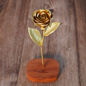 May include: A golden metal rose sculpture with detailed petals and leaves, mounted on a rectangular wooden base. The base has the text "Camella Bees" engraved on it. The rose is a decorative item.