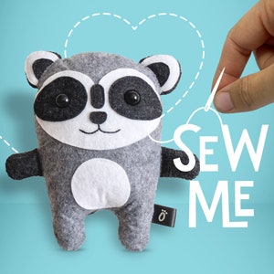 May include: A grey felt raccoon plush toy with black eyes and a white tummy. The toy is being held by a hand with a needle and thread, and the text "SEW ME" is written in white letters on a blue background.