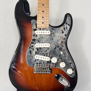 May include: An electric guitar with a sunburst finish body, a maple neck, and a distinctive grey and black marbled pickguard. The instrument features three white single-coil pickups, a chrome bridge, three control knobs, and a selector switch.