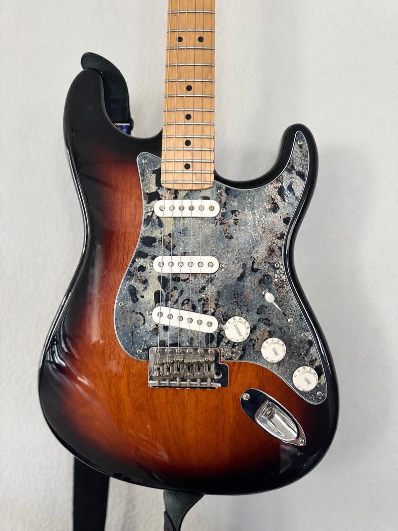 May include: An electric guitar with a sunburst finish body, a maple neck, and a distinctive grey and black marbled pickguard. The instrument features three white single-coil pickups, a chrome bridge, three control knobs, and a selector switch.