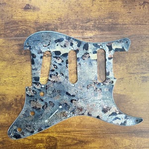 May include: A guitar pickguard featuring a mottled, multi-tonal pattern of grey, black, and metallic copper hues. The component includes three elongated cutouts for pickups, several smaller circular holes for mounting screws, and a volume knob.