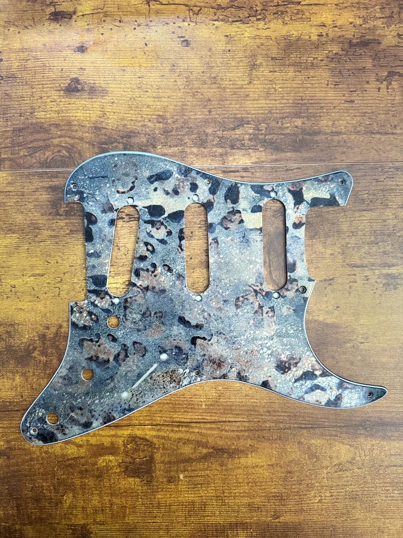 May include: A guitar pickguard featuring a mottled, multi-tonal pattern of grey, black, and metallic copper hues. The component includes three elongated cutouts for pickups, several smaller circular holes for mounting screws, and a volume knob.
