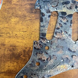 May include: A guitar pickguard featuring a textured, mottled pattern in shades of black, gray, and metallic gold. The component includes pre-cut openings for pickups and controls, along with small circular holes for mounting screws along the edges.