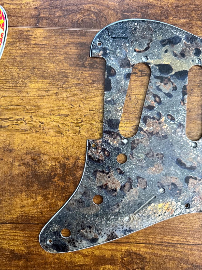 May include: A guitar pickguard featuring a textured, mottled pattern in shades of black, gray, and metallic gold. The component includes pre-cut openings for pickups and controls, along with small circular holes for mounting screws along the edges.