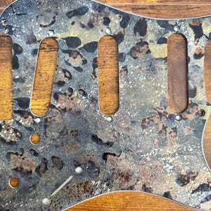 May include: A guitar pickguard featuring a mottled, multi-colored pattern of black, grey, gold, and copper tones. The component includes three elongated cutouts for pickups, several smaller circular holes for mounting screws, and a slot for a switch.