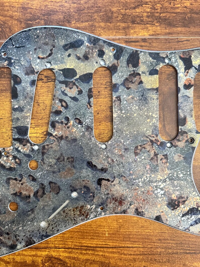 May include: A guitar pickguard featuring a mottled, multi-colored pattern of black, grey, gold, and copper tones. The component includes three elongated cutouts for pickups, several smaller circular holes for mounting screws, and a slot for a switch.