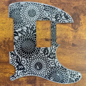 May include: A guitar pickguard featuring a silver base with a black floral pattern of large chrysanthemum-like flowers and swirling leaves. The pickguard has a rectangular cutout for a pickup and several small screw holes along the edges.