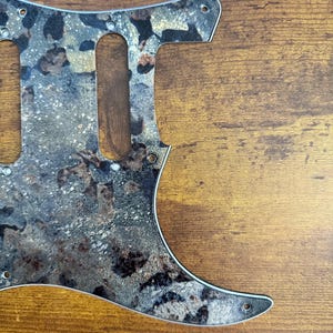 May include: A guitar pickguard featuring a mottled, textured pattern in shades of black, gray, brown, and metallic gold. The flat, shaped component includes pre-cut openings for pickups and multiple small circular holes for mounting screws.