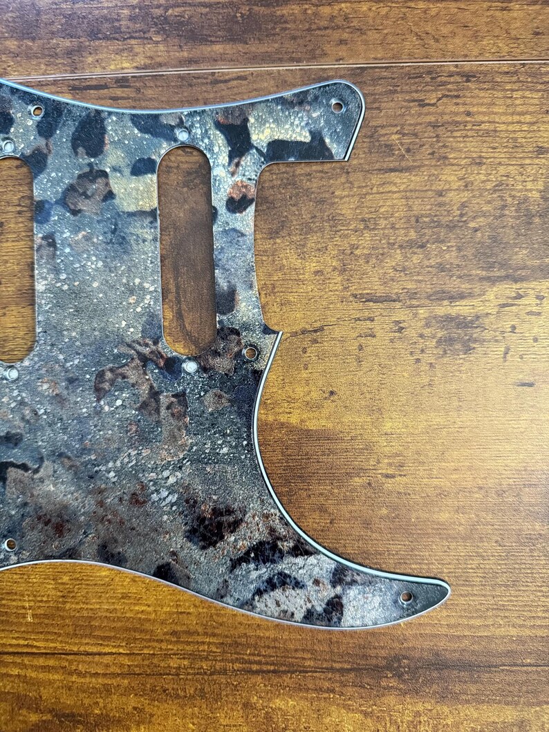 May include: A guitar pickguard featuring a mottled, textured pattern in shades of black, gray, brown, and metallic gold. The flat, shaped component includes pre-cut openings for pickups and multiple small circular holes for mounting screws.