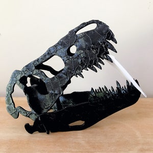 May include: Black and green dinosaur skull replica with a textured surface. The skull is open-mouthed with visible teeth.