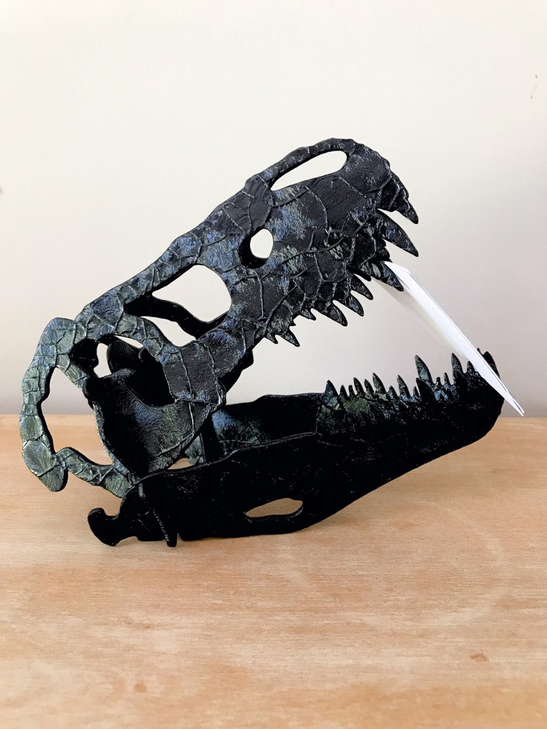 May include: Black and green dinosaur skull replica with a textured surface. The skull is open-mouthed with visible teeth.