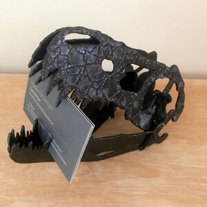 May include: Black 3D printed dinosaur skull business card holder. The holder is designed to hold business cards in the open mouth of the skull.