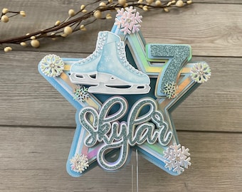 May include: A blue star-shaped cake topper with a pair of ice skates, the number 7, and the name "Skylar" in glitter lettering. The topper is decorated with snowflakes.