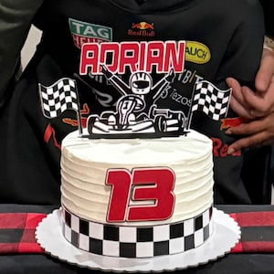 May include: A white frosted cake with a racing theme. The cake features a black and white checkered border, a red number 13, and a kart racing topper with the name "Adrian". Two checkered flags complete the design.