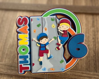 以下が含まれることがあります： 赤、黄、青の文字で「THOMAS」と書かれた紙製のケーキトッパー。カラフルなホールドが付いた灰色の岩壁を登る2人の漫画キャラクターが描かれています。デザインの右側には大きな青い数字の6が取り付けられています。