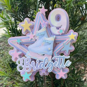 Könnte beinhalten: Ein mehrschichtiger Stern-Kuchentopper in Pastellrosa, Lila und Blau mit einem Paar Rollschuhen, der Zahl 9 und dem Namen Bridgette. Der Stern ist mit kleinen Sternen verziert.