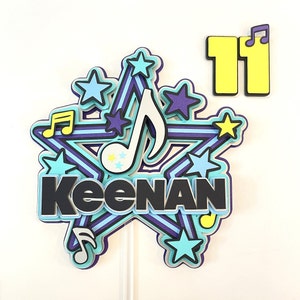 Puede incluir: Un adorno para tarta en capas con el nombre "Keenan" en letras negras, rodeado de un diseño de estrella con notas musicales y estrellas en tonos azules, morados y amarillos. También se incluye el número "11".