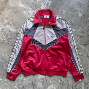 Puede incluir: Chaqueta deportiva Kappa roja, gris y plateada con cremallera completa. La chaqueta presenta un cuello alzado, el logotipo de Kappa en el pecho y rayas blancas con el logotipo de Kappa en las mangas. La chaqueta es de material brillante.
