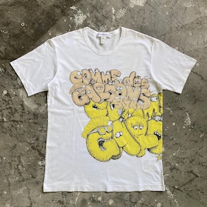 Puede incluir: Camiseta blanca con un diseño gráfico amarillo y marrón. El diseño incluye el texto "Comme des Garcons" en un estilo de dibujos animados. La camiseta tiene cuello redondo y mangas cortas.