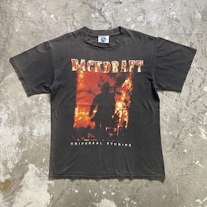 Può includere: T-shirt grigio scuro con il titolo del film "BACKDRAFT" in lettere rosse e arancioni sopra una silhouette di una persona davanti alle fiamme. Il logo Universal Studios è visibile nella parte superiore e inferiore della maglietta.