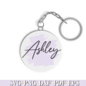 Pode incluir: Um chaveiro circular branco com um fundo de aquarela roxo claro e o nome "Ashley" escrito em it&aacute;lico preto.