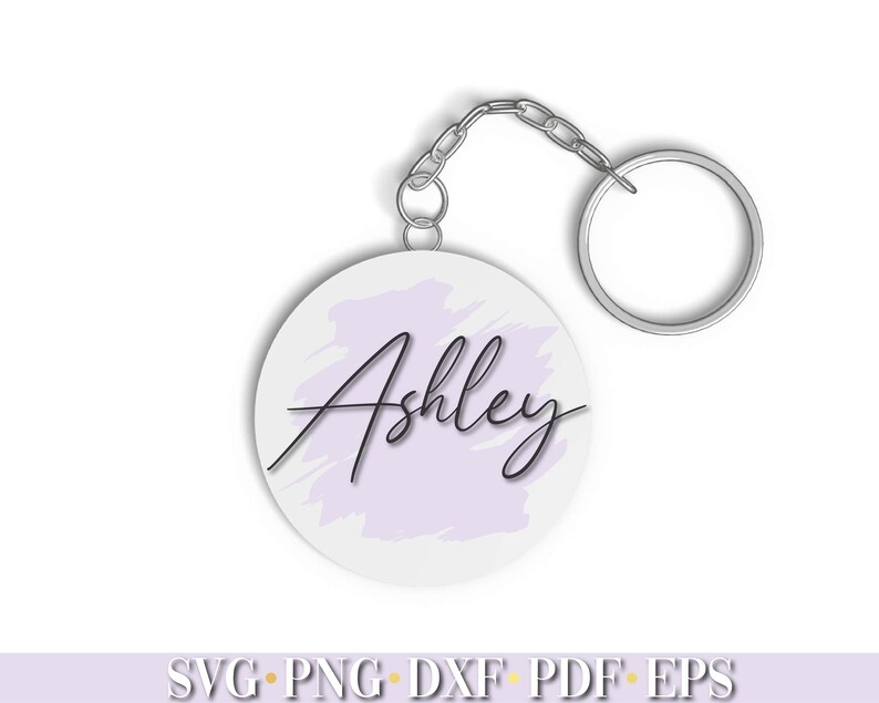 Pode incluir: Um chaveiro circular branco com um fundo de aquarela roxo claro e o nome "Ashley" escrito em it&aacute;lico preto.