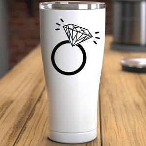 Pode incluir: Um copo de a&ccedil;o inoxid&aacute;vel branco com um design de anel de diamante preto. O anel tem um diamante grande com um efeito brilhante.