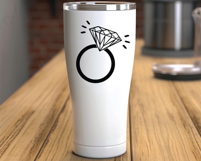 Pode incluir: Um copo de a&ccedil;o inoxid&aacute;vel branco com um design de anel de diamante preto. O anel tem um diamante grande com um efeito brilhante.
