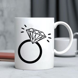 Pode incluir: Caneca de cer&acirc;mica branca com um gr&aacute;fico preto de um anel de diamante. O anel &eacute; estilizado com um design simples e o diamante &eacute; delineado com linhas pretas.