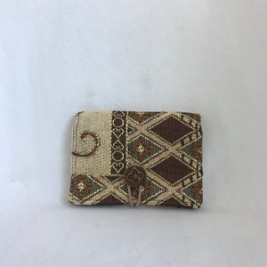May include: A rectangular wallet with a beige and brown geometric pattern and a button closure. The design includes diamond and floral motifs. The button is copper-coloured, and the wallet is secured with a beige string.