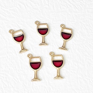 May include: Five gold-colored wine glass charms with a deep red enamel filling. Each charm has a small loop at the top for attaching to jewelry or other crafts. The wine glasses are a simple, elegant design.