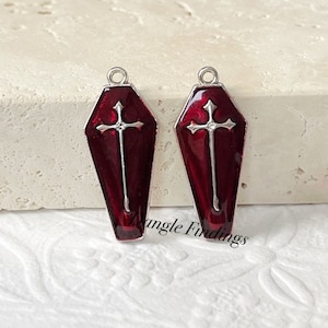 May include: Two silver-toned coffin-shaped charms with a red enamel finish and a silver cross design. The charms have a loop at the top for attaching to jewelry.