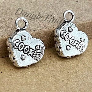 May include: Two silver heart-shaped charms with the word "COOKIE" embossed on them. Each charm has a loop at the top for attaching to a necklace or bracelet.