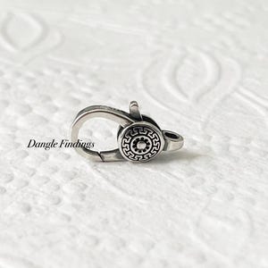 May include: A silver-toned jewelry clasp with a circular decorative element. The clasp has a spring-loaded mechanism and a decorative circular design with a repeating pattern. The text "Dangle Findings" is visible on the left side.