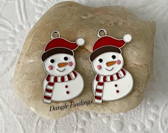 May include: Two silver snowman charms with red hats, white bodies, and red and white striped scarves. Dangle Findings is printed on the rock they are sitting on.