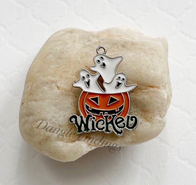 Pode incluir: Um amuleto de prata com uma ab&oacute;bora e tr&ecirc;s fantasmas. A ab&oacute;bora &eacute; laranja com uma cara preta e os fantasmas s&atilde;o brancos. O amuleto diz "Wicked" em letras prateadas com acentos verdes.