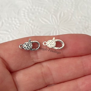 May include: Two small, silver-colored lobster claw clasps. Each clasp features an ornate, swirling design on the rounded end. The clasps are shown against a soft, textured background.