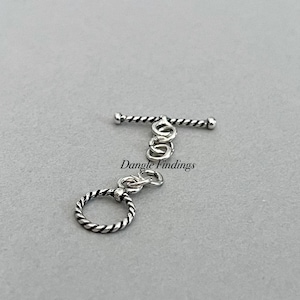 May include: Silver toggle clasp with a twisted rope design. The clasp has a round ring on one end and a bar with two small balls on the other end.