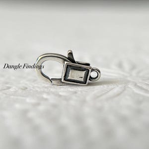 May include: A silver-colored lobster clasp with a rectangular design and a small loop. The clasp is made of metal and has the text "Dangle Findings" printed on the left side.