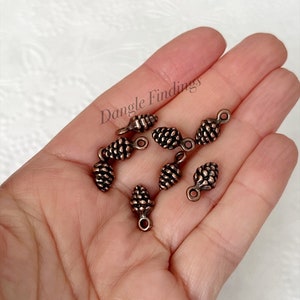 May include: A close-up of eight copper-colored pine cone charms with a loop at the top. The charms are arranged in a hand, with the text "Dangle Findings" visible in the background.