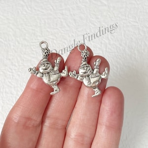 May include: Two silver-toned snowman charms with a snowflake on top of their hats. The snowman charms are in a skating pose with their arms outstretched.