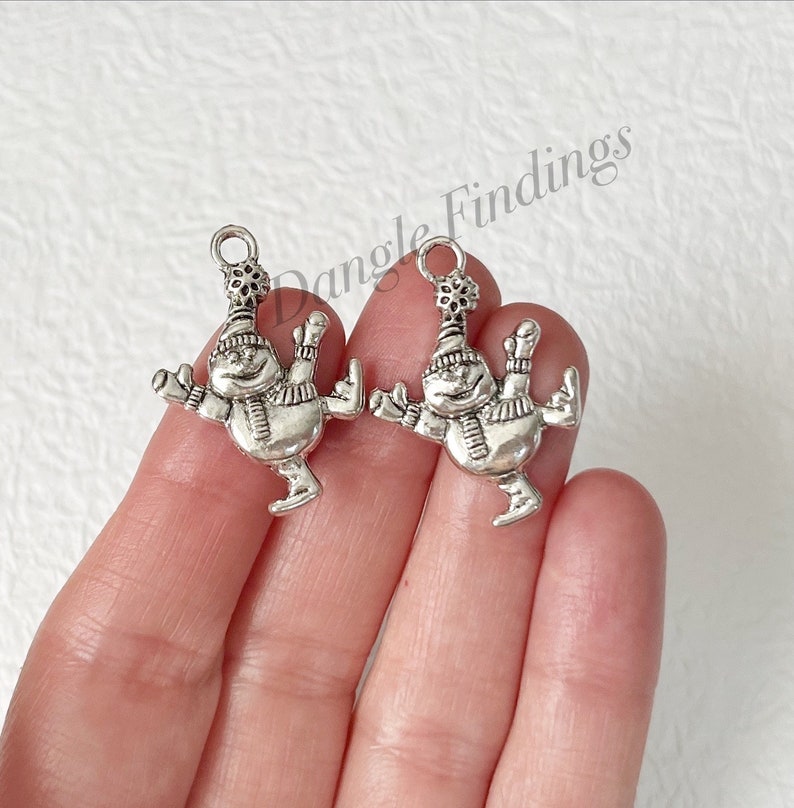 May include: Two silver-toned snowman charms with a snowflake on top of their hats. The snowman charms are in a skating pose with their arms outstretched.