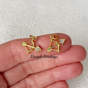 May include: Two gold-colored charms, each featuring a bow and arrow design with a triangle-shaped arrow head. The arrowheads are encrusted with small, clear crystals. The charms are shown on a light background. Text on the image reads "Dangle Findings".