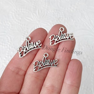 May include: Three silver-colored charms with the word "Believe" in a cursive font. Each charm has a small loop at the top for attaching to jewelry or other crafts. The charms are held in a hand.