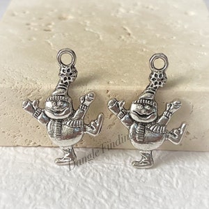 May include: Two silver charm pendants shaped like a snowman wearing a hat and scarf, ice skating. The snowman has a round face with a smile and a small black dot for a nose.