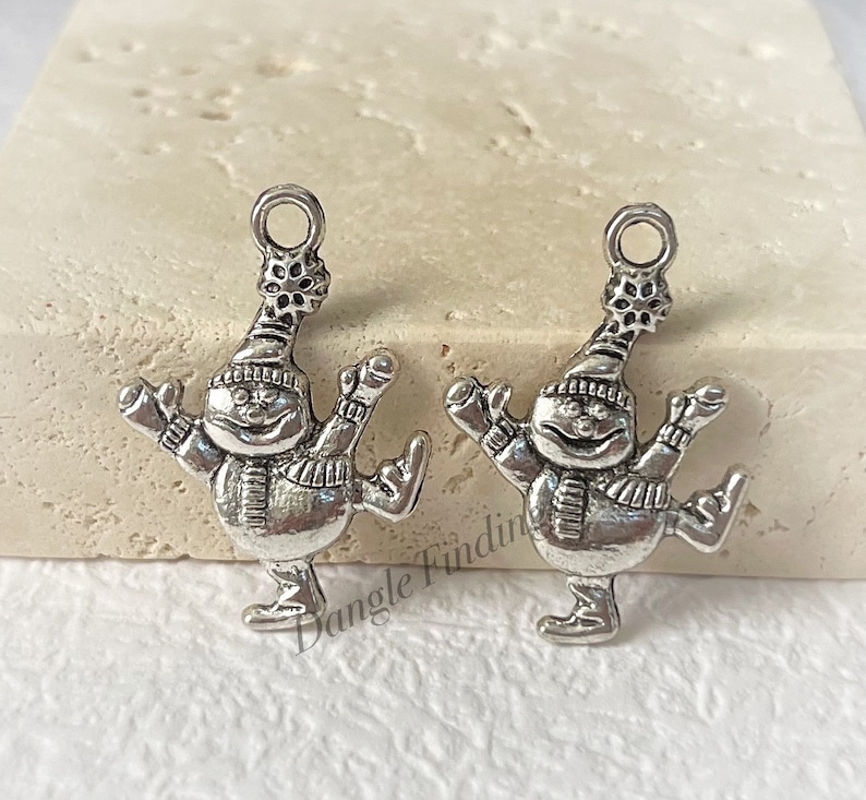 May include: Two silver charm pendants shaped like a snowman wearing a hat and scarf, ice skating. The snowman has a round face with a smile and a small black dot for a nose.