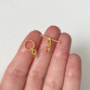 May include: Gold-toned metal toggle clasp with a twisted rope design. The clasp has a round ring on one end and a bar with three connecting rings on the other end.