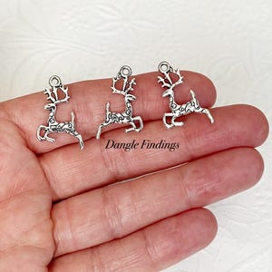May include: Silver-toned reindeer charms with intricate detailing. The charms feature a running reindeer design with antlers and decorative patterns. The text "Dangle Findings" is visible. These charms are ideal for jewelry making and crafting projects.